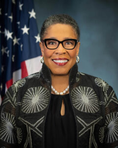 Marcia L. Fudge stands in front of an American flag and a blue background, wearing black glasses