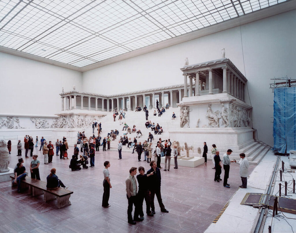 A large museum gallery with an architectural reproduction of a classical building. The gallery is filled with many people and has a roof with many window.