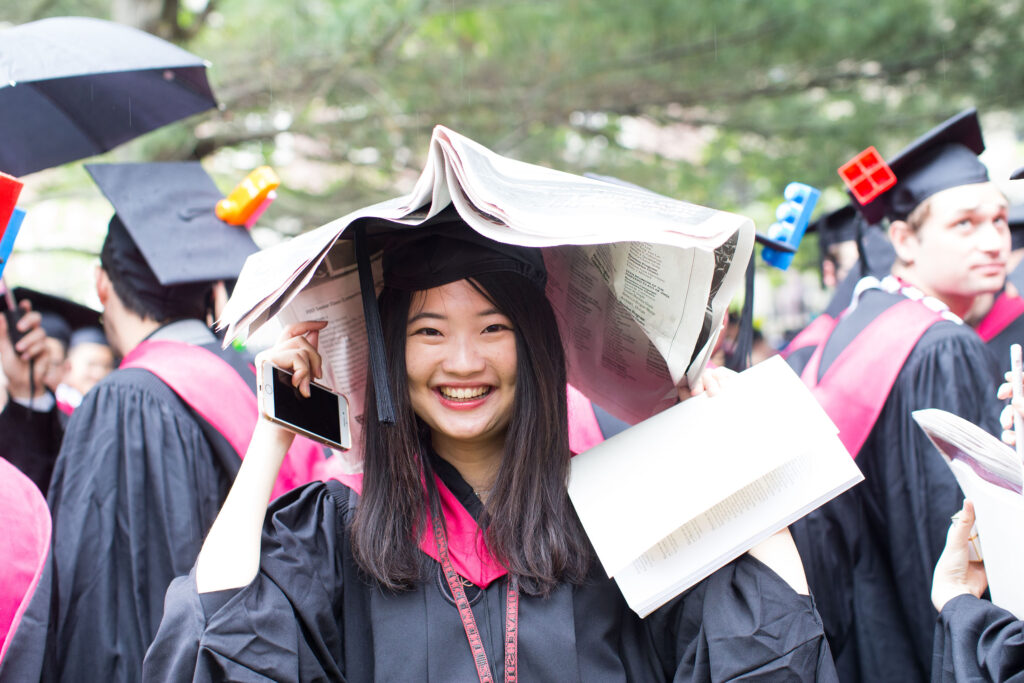 A student wearing Harvard cap and gown grins to the camera while covering her head with a newspaper to protect herself from the rain. 