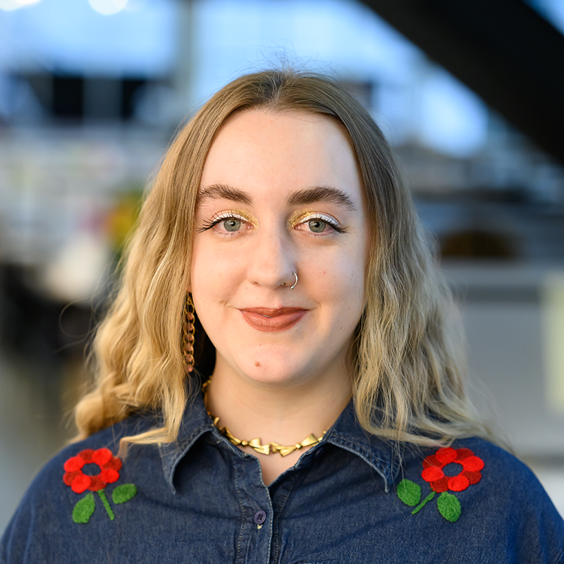 Mila Cuda stands in front of trays in blue denim button up shirt with red flower embroidery on the shoulders