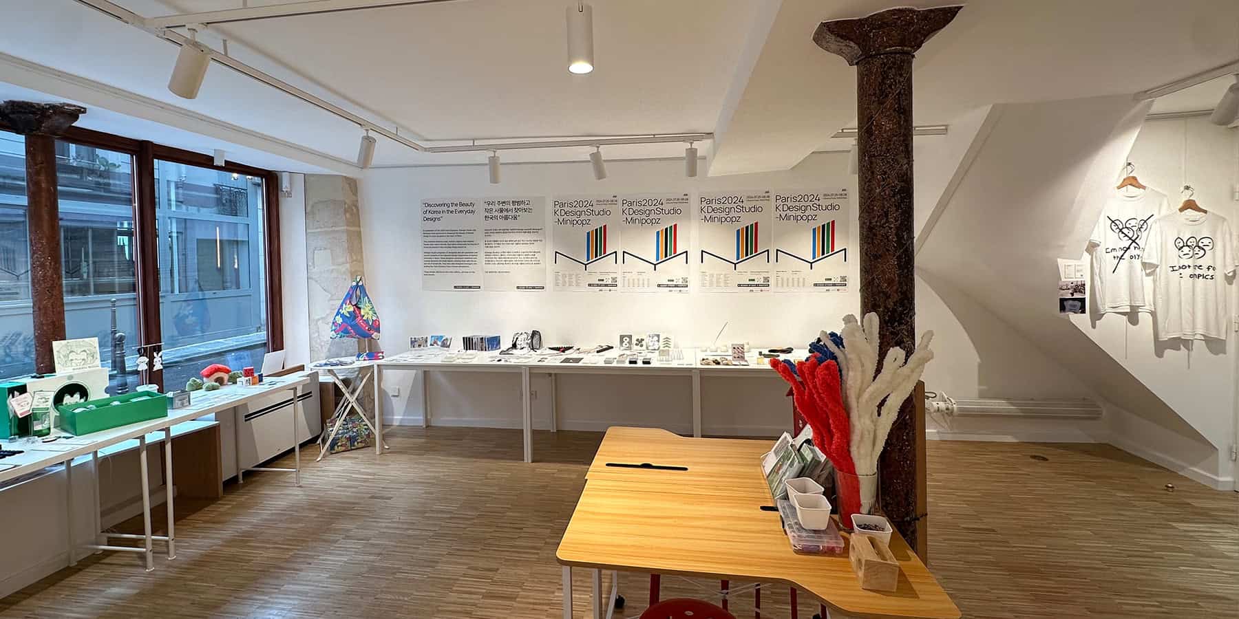 KDesign Studio exhibition, featuring white walls with colorful posters, Olympic t shirts, and tables displaying various objects, such as bags, incense holders, and character drawings.