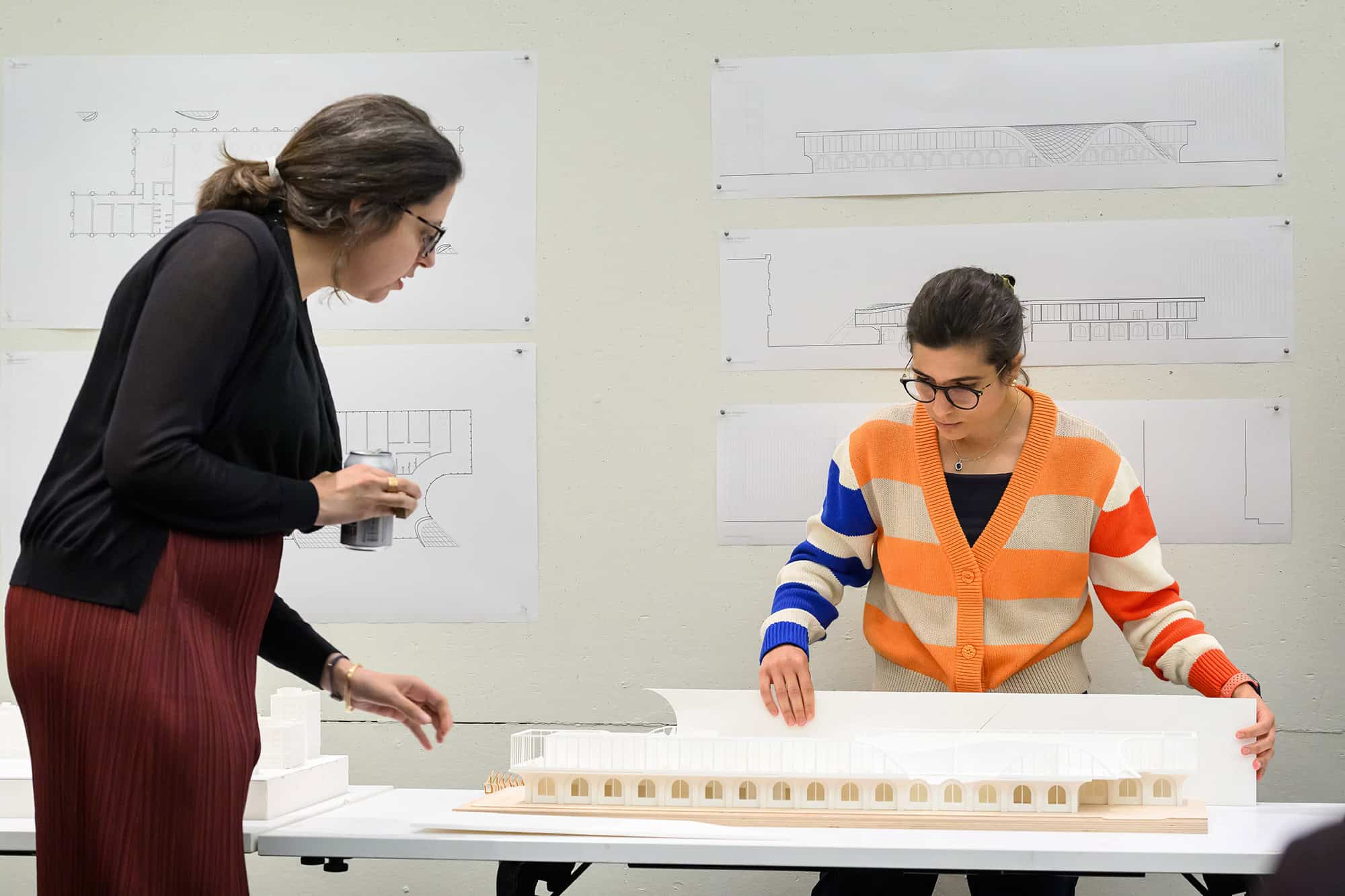 Two students working on architectural model with sketches in the background