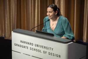 Miriam Hernandez Medina in a teal dress speaks at the Harvard GSD podium